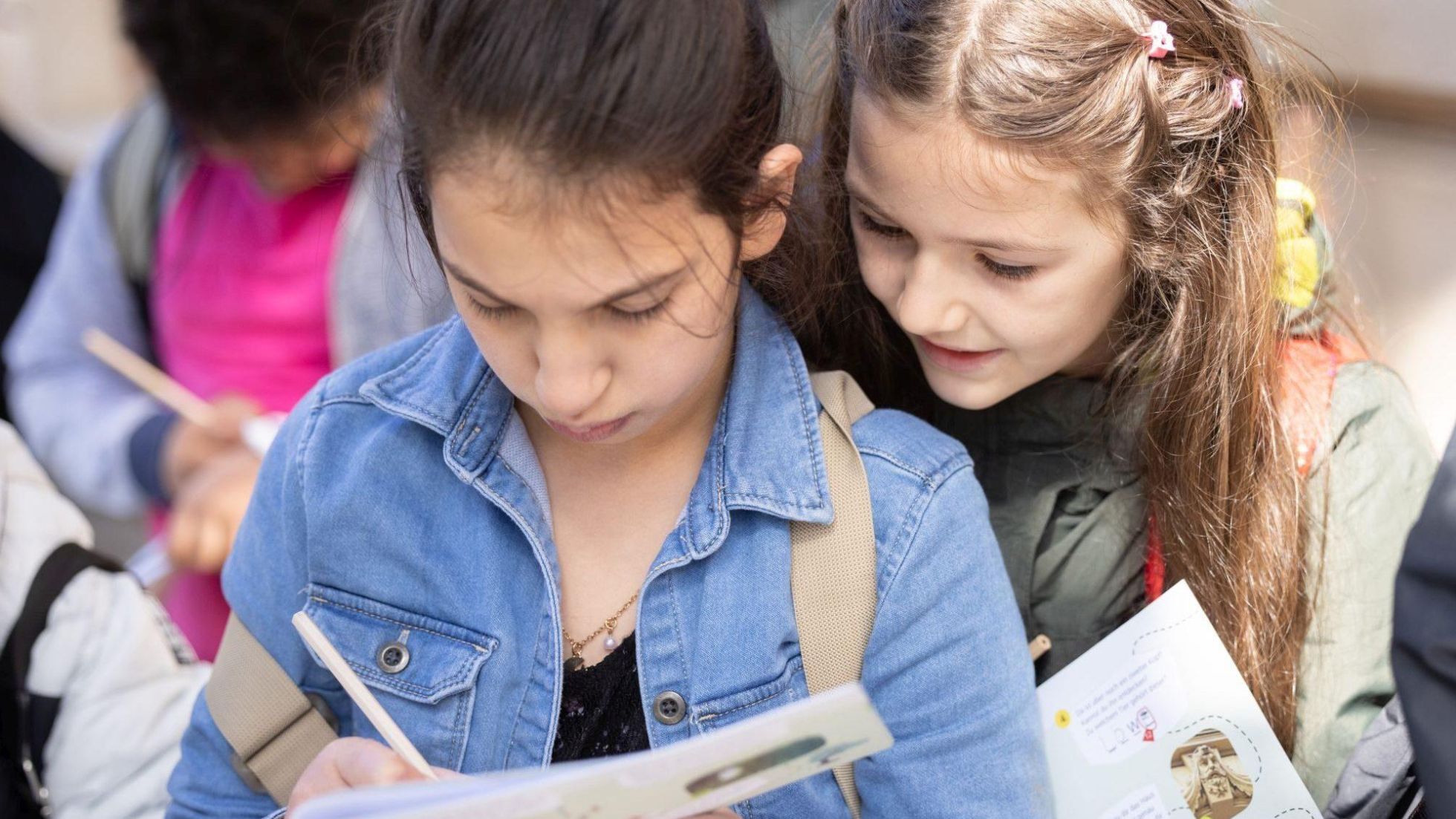 Ein Mädchen macht Notizen in einem Heft, ein anderes Mädchen schaut ihr dabei über die Schulter.