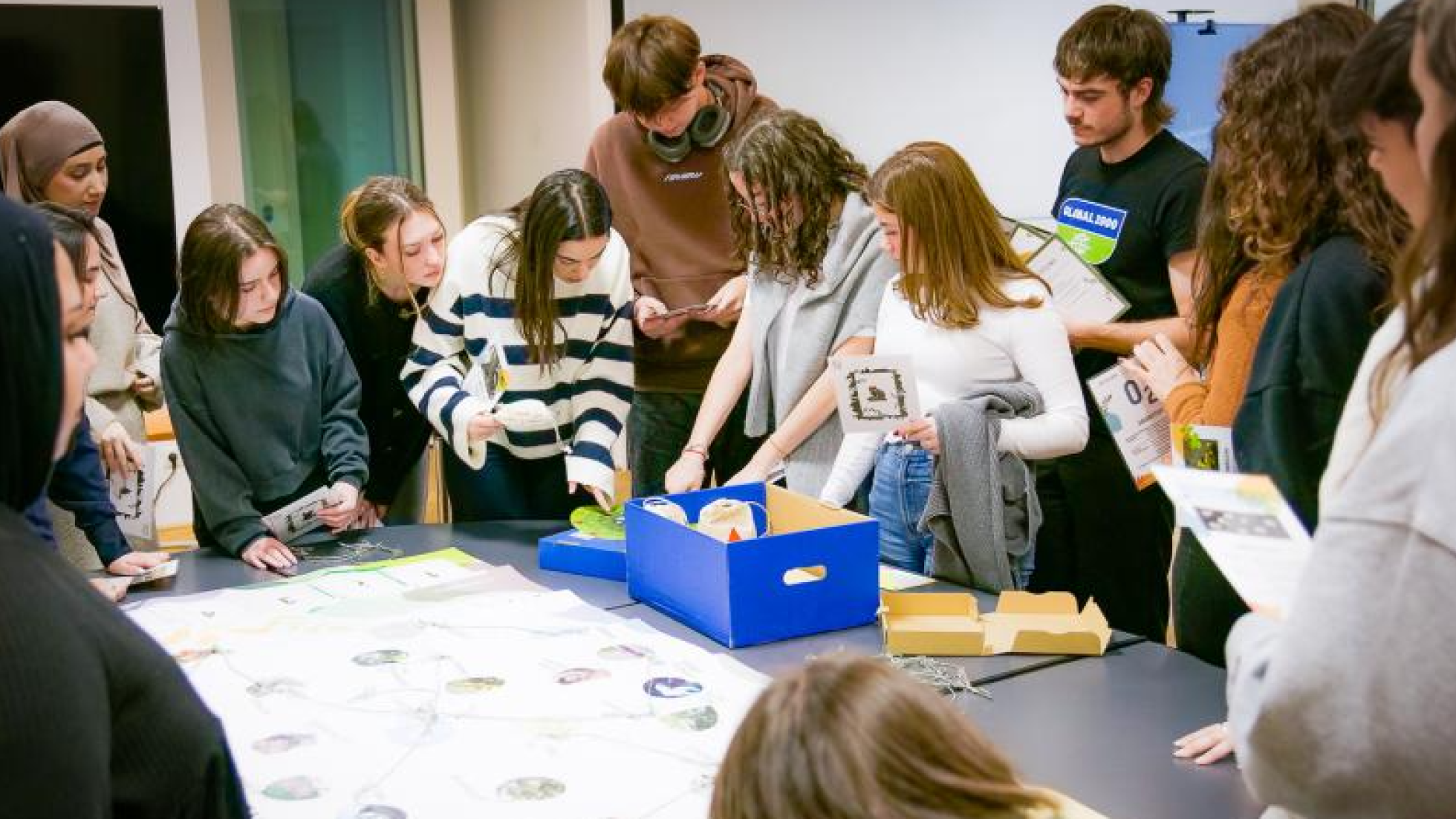 Eine Gruppe Jugendlicher steht um einen Tisch herum. Auf dem Tisch liegt ein Plakat und eine blaue Schachtel.