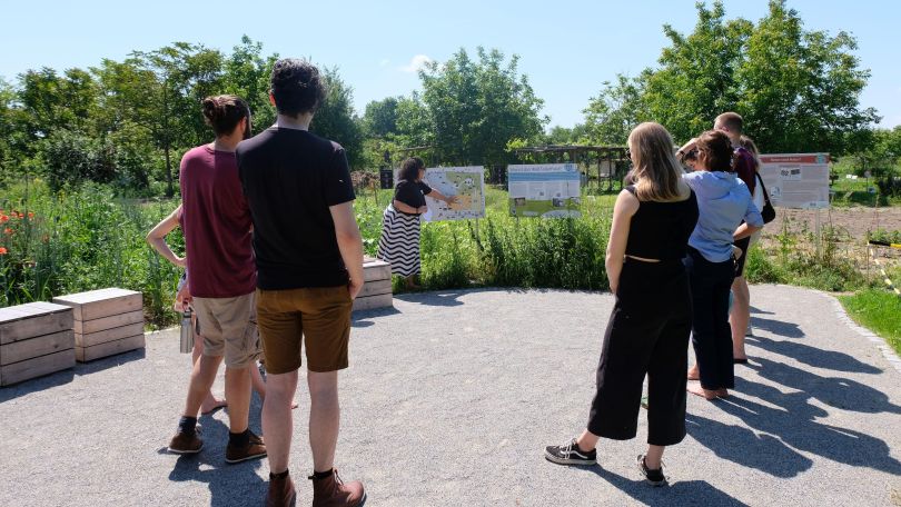 copyright: WeltTellerFeld Eine Gruppe Erwachsener steht draußen im Grünen im Halbkreis vor einer Schautafel. Eine junge Frau erklärt etwas an der Tafel.