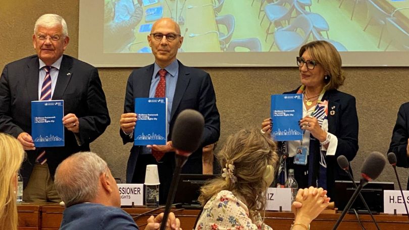 copyright: Shams Asadi 3 Vortragende halten die UN-Leitlinien in Druckform vor Publikum, im Hintergurnd eine Leinwand mit Präsentation