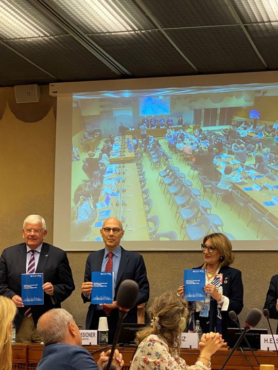 copyright: Shams Asadi 3 Vortragende halten die UN-Leitlinien in Druckform vor Publikum, im Hintergurnd eine Leinwand mit Präsentation