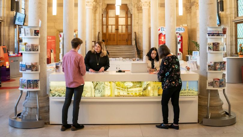 copyright: Stadt Wien - David Bohmann An einem Infoschalter im Rathaus beraten Mitarbeiterinnen 2 Besucher*innen.