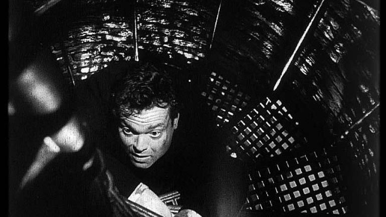 Black-and-white film still: Orson Welles on a spiral staircase
