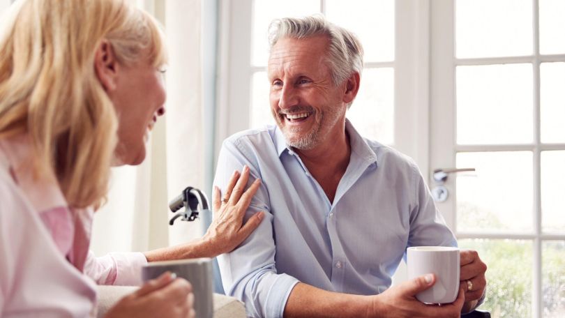 Ein Mann und eine Frau mittleren Alters trinken gemeinsam Tee und lachen herzlich. Der Mann sitzt im Rollstuhl und trägt ein schickes, helles Hemd.