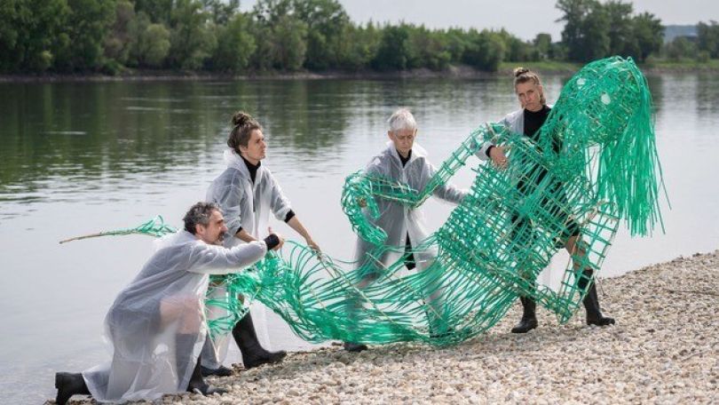 copyright: Michael Gizicki 4 Menschen ziehen eine grüne Meerjungfrauen-Skulptur aus einem Gewässer. Alle tragen schwarze Kleidung und darüber einen weißen, transparenten Umhang aus Kunststoff.