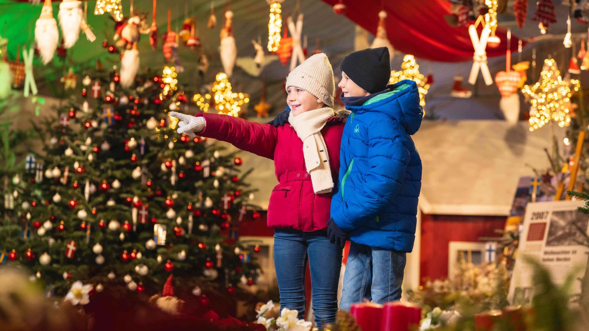 2 Kinder in Winterjacken stehen zwischen Weihnachtsdeko und einem Christbaum.