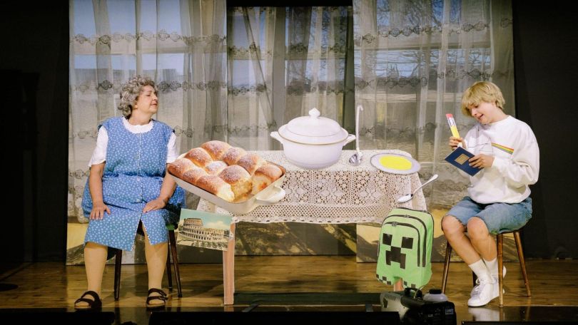 Oma und Junge sitzen sich an einem Tisch gegenüber. Es sind Buchteln, ein Topf und andere Dinge zu sehen.