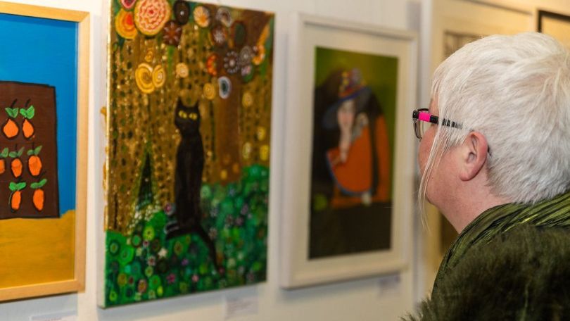 Eine Frau mit kurzen grauen Haaren betrachtet ein Gemälde von einer Katze. Daneben hängen noch mehr Bilder an der Wand.