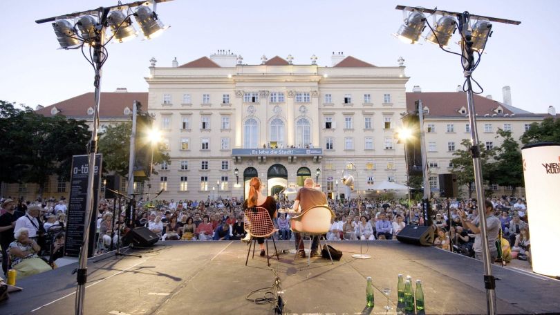 copyright: Simon Brugner 2 Personen im Freien auf einer Bühne, dahinter viele Menschen, die zuhören
