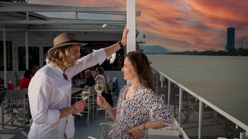 Zwei Personen an Board des Schiffes MS Kaiserin Elisabeth auf der Donau. Sie lachen einander an und haben Sektgläser in der Hand. Im Hintergrund ein rosa-blauer Sonnenuntergang.