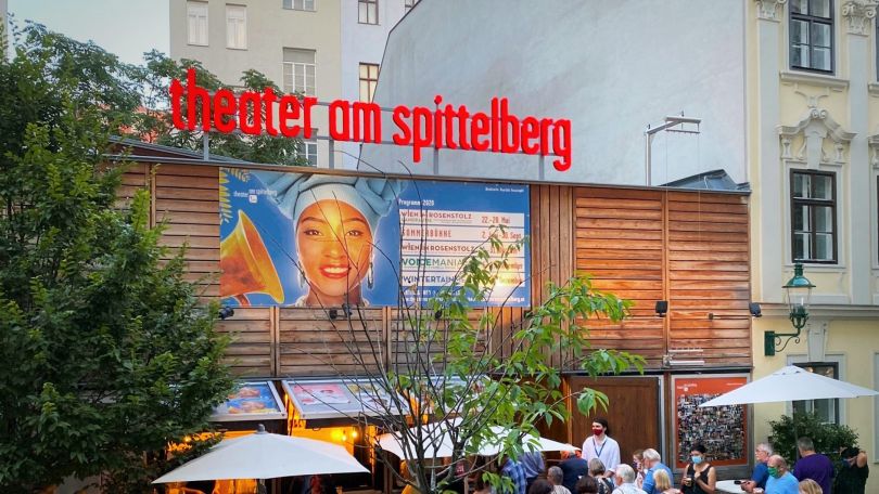 copyright: Theater am Spittelberg Archiv Außenansicht des Theaters, großer, roter Schriftzug über dem Dach, auf der Wand ein großes Poster mit einer Frau und dem Spielplan. Vor dem Theater stehen einige Menschen und gehen hinein.