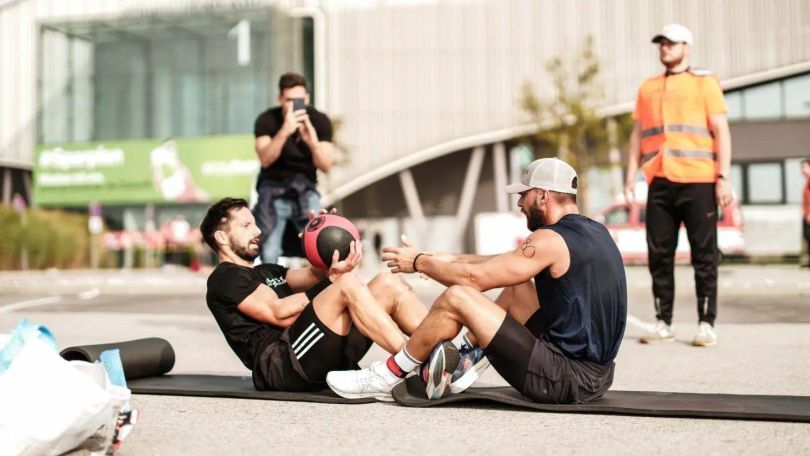 copyright: FITLETIX Zwei Männer machen draußen auf einer Sportmatte Situps mit einem Gymnastikball. Im Hintergrund ein Fotograf und eine Security-Person.