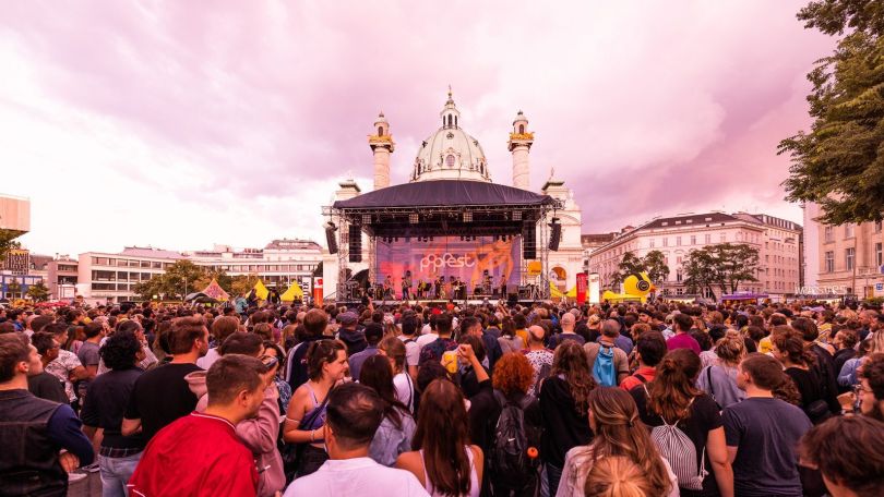 Zentrale Sicht auf eine Bühne vor der Karlskirche mit Banner "Popfest", davor eine dichte Menge an Menschen. Der Himmel ist mit rosa Wolken bedeckt.