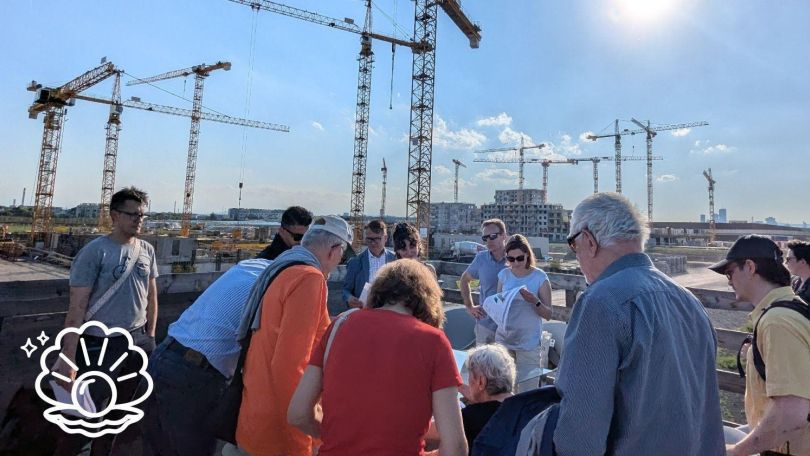 copyright: GB* Ungefähr 15 Personen stehen vor einer Baustelle und schauen sich Stadtpläne an. Im Hintergrund sind mehrere Kräne zu sehen.