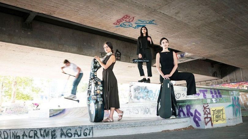 Drei weiblich gelesene Personen in schwarzer Kleidung stehen unter einer U-Bahn-Brücke mit Parkour-Anlage und posieren mit ihren Instrumenten für die Kamera. Im Hintergrund ein verschwommene Person, die mit ihrem Roller Tricks macht.