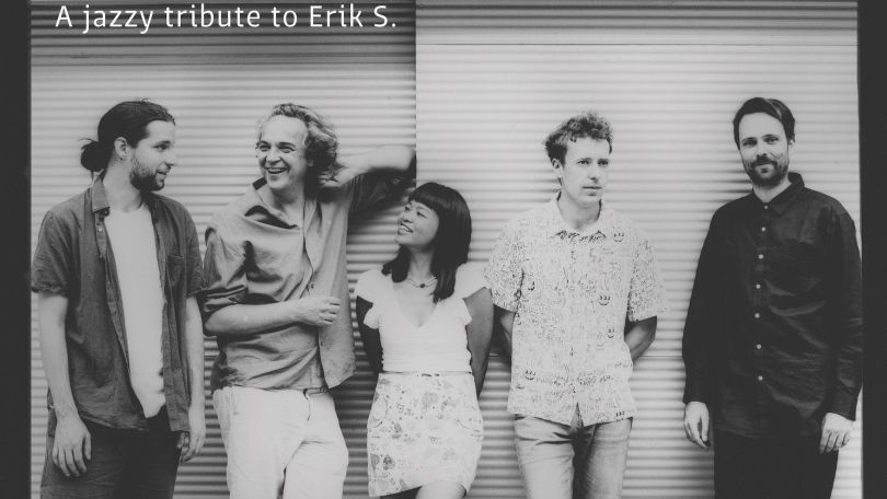 copyright: Kulturverein dusha connection Schwarz-weiß Aufnahme einer fünfköpfigen Gruppe, vor einer schlichten gestreiften Wand. Die ersten drei Menschen von links reden und lachen miteinander. Oben links Text "A jazzy tribute to Erik S."