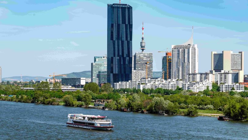 copyright: DDSG Blue Danube Ausflugsschiff fährt auf der Donau, im Hintergrund hellblauer Himmel und hohe Gebäudekomplexe.