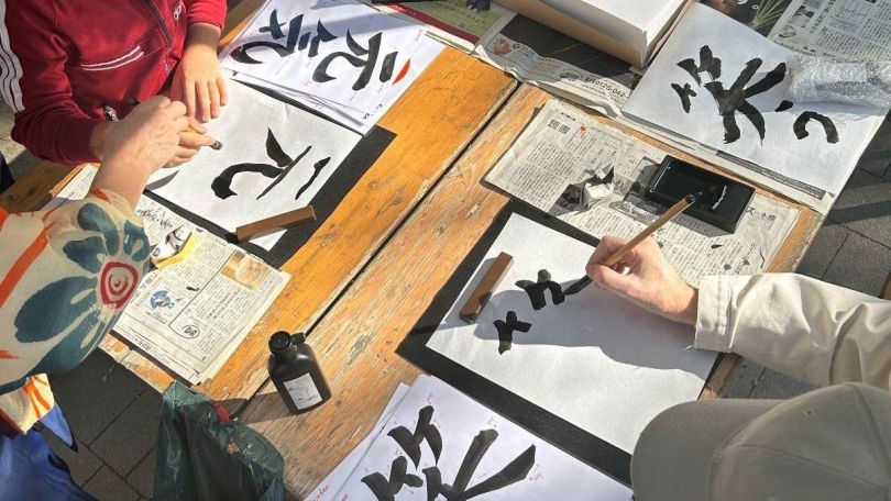 copyright: BV 16 Ansicht von oben auf einen Holztisch im Freien an dem zwei Personen sich mit japanischer Kalligraphie beschäftigen. Mehrere Blätter Papier, alte Zeitungen als Unterlagen für die Tinte.
