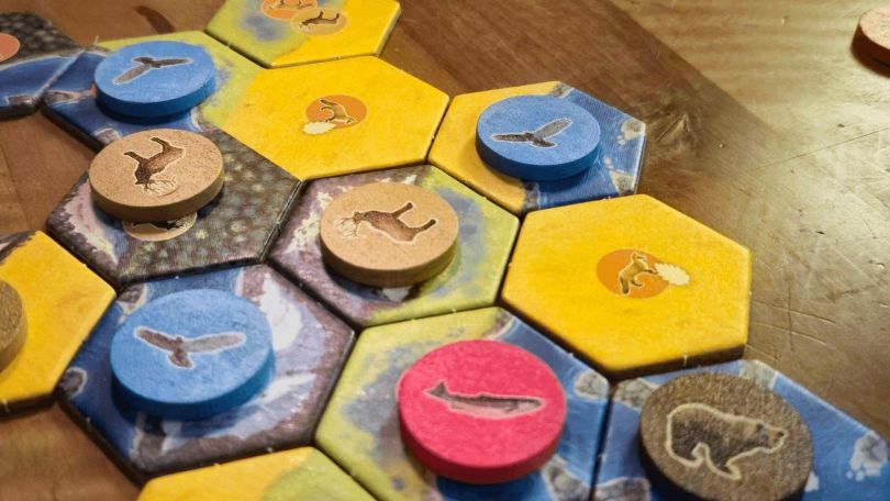 Nahaufnahme eines Brettspiels auf einem Holztisch. Sechseckige Spielplättchen mit verschiedenen Landschaftsillustrationen liegen aneinander und auf manchen sind runde Spielsteine mit Tierillustrationen drauf,