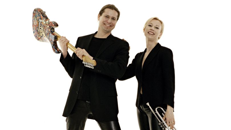 copyright: Christian Schreibmüller Zwei Personen in schwarz gekleidet vor einem weißen Hintergrund lächeln in die Kamera. Die Person links hat die braunen Haare nach hinten in einen Zopf gebunden und hält eine E-Gitarre am Griff wie eine Axt. Die Person rechts hat die blonden Haare nach hinten in einen Zopf gebunden und hält eine Trompete und hält die Person an der Schulter.