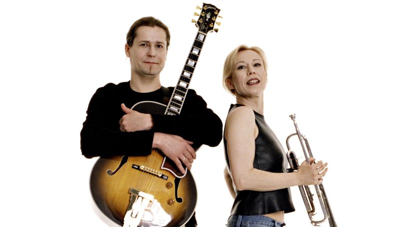 copyright: Christian Schreibmüller Eine Person mit langärmligem schwarzem Oberteil steht frontal zur Kamera und hält eine Gitarre in den Armen. Eine Person mit ärmellosem schwarzem Oberteil steht seitlich zur Kamera und hält eine Trompete in der Hand. Die beiden stehen vor einem weißen Hintergrund.