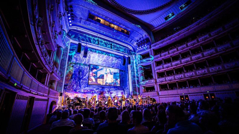 copyright: Rahel Gölzner Bühne in der Wiener Staatsoper in dunkler blau-lilanen Beleuchtung. Auf der Bühne ist ein Orchester mit Sänger*innen zu sehen und ein Bildschirm zeigt die Bühne in nah für die hinteren Plätze.