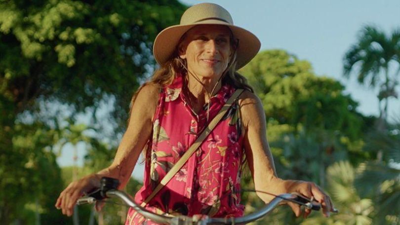 Person mit Sonnenhut und pinkem ärmellosen Kleid mit Blumenmuster, fährt mit dem Fahrrad und lächelt leicht, im Hintergrund verschwommene Bäume und ein hellblauer Himmel.