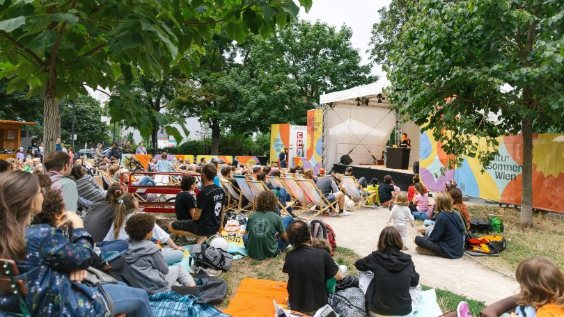 copyright: Theresa Wey Zuseher*innen auf Matten und Bänken vor einer Freiluftbühne in einem Park