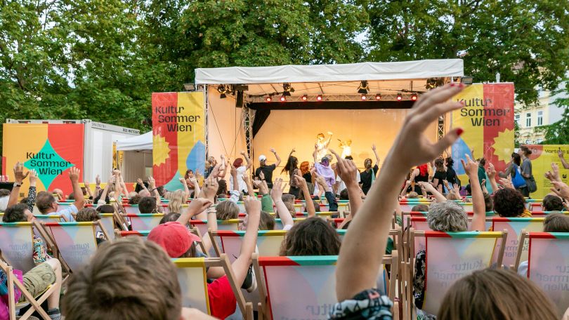 copyright: Judith Stehlik Bühne mit drei bunten "Kultursommer Wien" Bannern. Auf der Bühne stehen zwei Personen mit Mikrofon und heben eine Hand in die Luft, das Publikum macht mit. Vorne stehen Erwachsene, hinten sitzen auch Menschen in Liegestühlen.