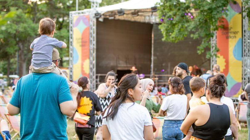 Zuseher*innen vor einer Freiluftbühne inmitten von Parkbäumen