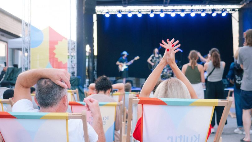 copyright: Paola Lesslhummer Eine blau beleuchtete Bühne mit Musiker*innen mit Gitarren, davor ein Publikum welches teils steht und teils in Liegestühlen sitzt. Ganz nah sind zwei sitzende Personen zu sehen, von denen eine mit den Händen über dem Kopf klatscht.