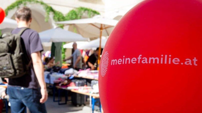 copyright: Erzdiözese Wien/R.Hafellner Im Vordergrund eine roter Luftballon mit Aufdruck "meinefamilie.at", im Hintergrund verschwommen Bänke, verdeckt von Sonnenschirmen und einige Menschen.