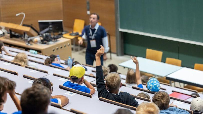 copyright: Kinderbüro der Universität Wien/APA-Fotoservice/Juhasz Innenansicht eines Hörsaals, vorne steht eine Person und erzählt etwas, in den Reihen sitzen lauter Kinder, manche schreiben mit, manche zeigen auf.