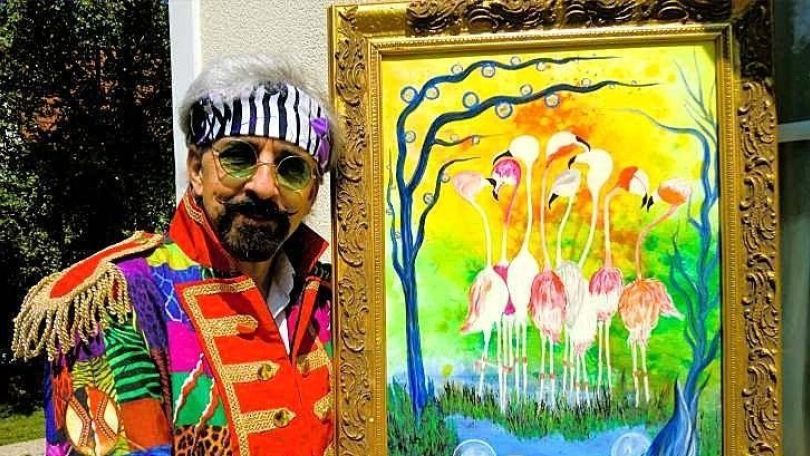 copyright: Robin Künstler Bunt gekleideter Mann mit grüner Sonnenbrille und Schnauzbart posiert mit einem gold eingerahmten Gemälde von Flamingos an einem Teich