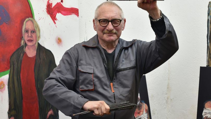 copyright: kulturimdritten Mann mit Brille und rasierten Haaren schaut in die Kamera und hebt die Faust, Hinter ihm ein Gemälde einer Frau in einem roten Kleid und schwarzem Mantel.