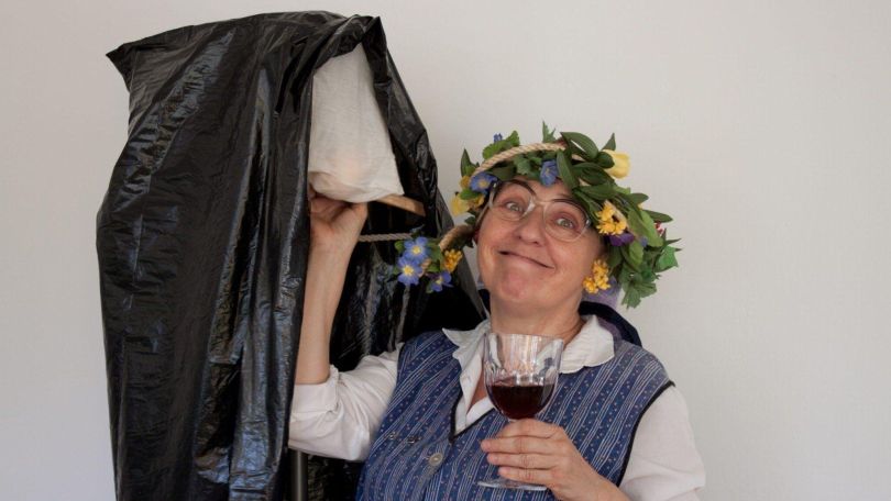 copyright: Theater-Center-Forum Lächelnde Person mit Blumenkranz hebt einen Kleiderständer mit Müllsack und hält ein Glas Rotwein.