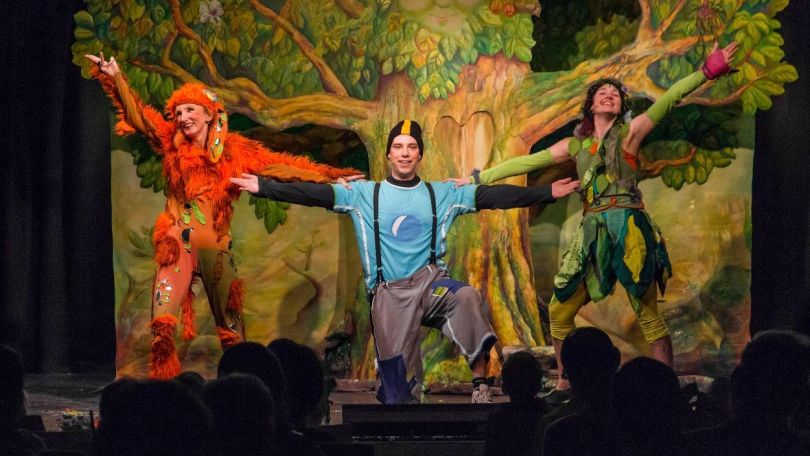 copyright: Theater Heuschreck 3 Schauspielerinnen in bunter Kleidung auf einer Bühne mit einer Wald-Kulisse als Hintergrund.