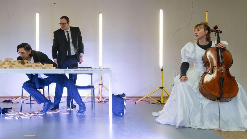 copyright: Teresa Thomaschütz Im Vordergrund sitzt eine Person in einem Hochzeitskleid auf einem Stuhl und hält ein Cello. Rechts und links hinter ihr stehen senkrechte Lichtstäbe auf Stativen. Links im Bild sitzen und stehen zwei Personen in dunklen Anzügen an einem weißen Tisch, auf dem Holzklötze verteilt sind; einige Klötze liegen auf dem Boden.