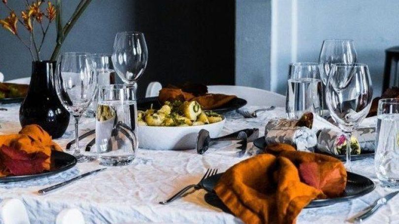 copyright: www.pexels.com / Rachel Claire Esstisch mit weißer Tischdecke. weißblauen Tellern und orangenen Stoffservietten, leere Weingläser und eine dunkelblaue Vase mit gelben Blumen.