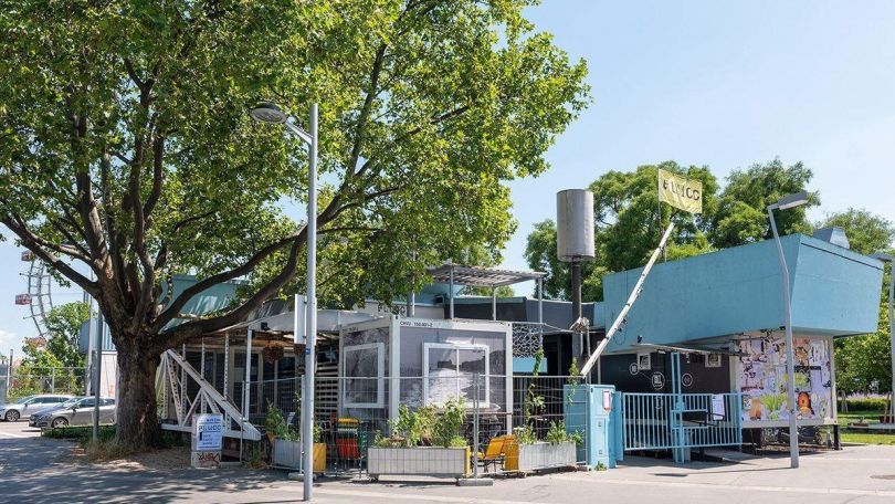 copyright: ola_queen Außenansicht auf ein kleines Gebäude mit vielen Zäunen und vielen Ebenen, Schilder sagen "FLUCC", daneben ein großer Baum, im Hintergrund das Riesenrad.