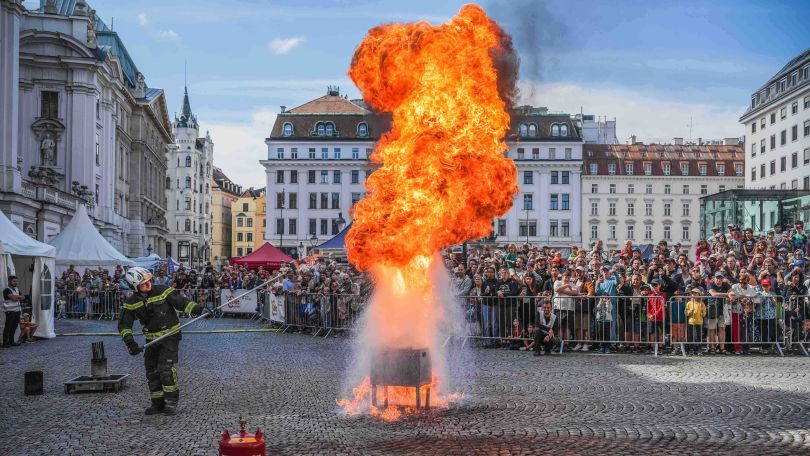 copyright: Stadt Wien – Feuerwehr Ein Feuerwehrmann löscht beim Feuerwehrfest einen riesigen Feuerball auf einem Platz in der Innenstadt. Hinter Absperrungen verfolgen viele Zuseher die Vorführung.