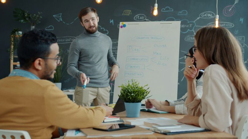 copyright: Hallo Klima! Ein Mann steht bei einem Meeting neben einem Flipchart. 3 Kolleg*innen machen sich an einem Besprechungstisch Notizen und hören zu.