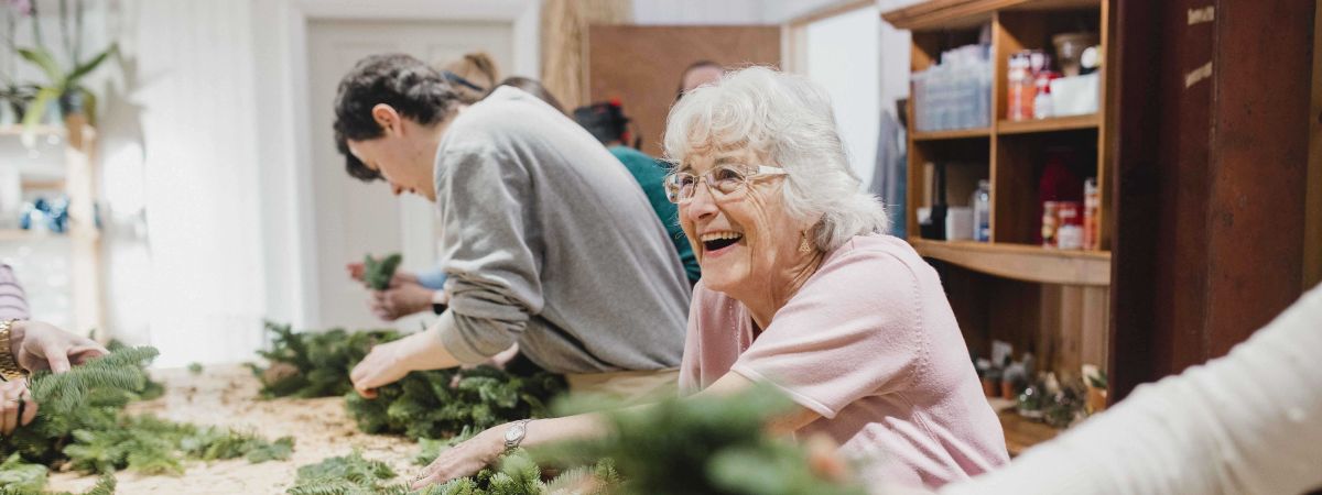 Eine ältere Frau mit weißen Haaren bastelt gemeinsam mit anderen Menschen an einem großen Holztisch mit Tannenzweigen.