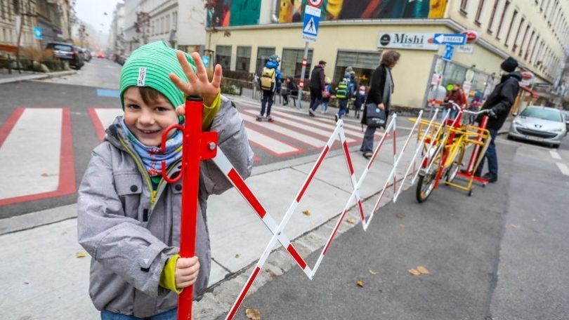 Ein Kind lächelt in die Kamera und hält sich an einer Straßenabsperrung fest. Im Hintergrund weitere Kinder mit Rucksäcken.
