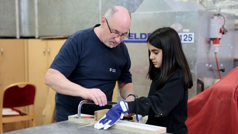 Ein Mädchen sägt ein Stück Holz. Ein Techniker hilft ihm.