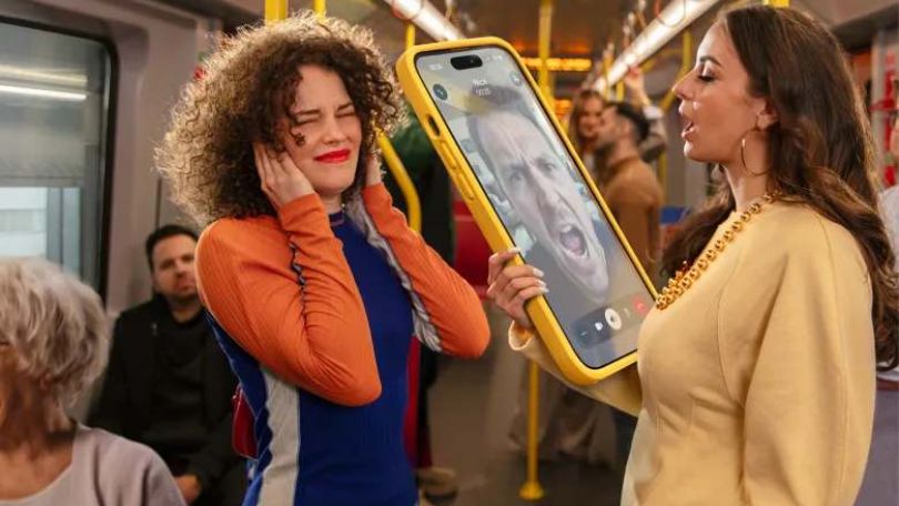 Eine Frau videotelefoniert mit einem übergroßen Telefon auf Lautsprecher in der U-Bahn. Die Frau neben ihr hält sich genervt die Ohren zu.