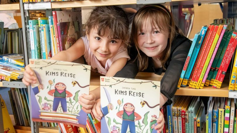2 Kinder strecken ihren Kopf durch ein Bücherregal heraus und halten jeweils 1 Kinderbuch in den Händen