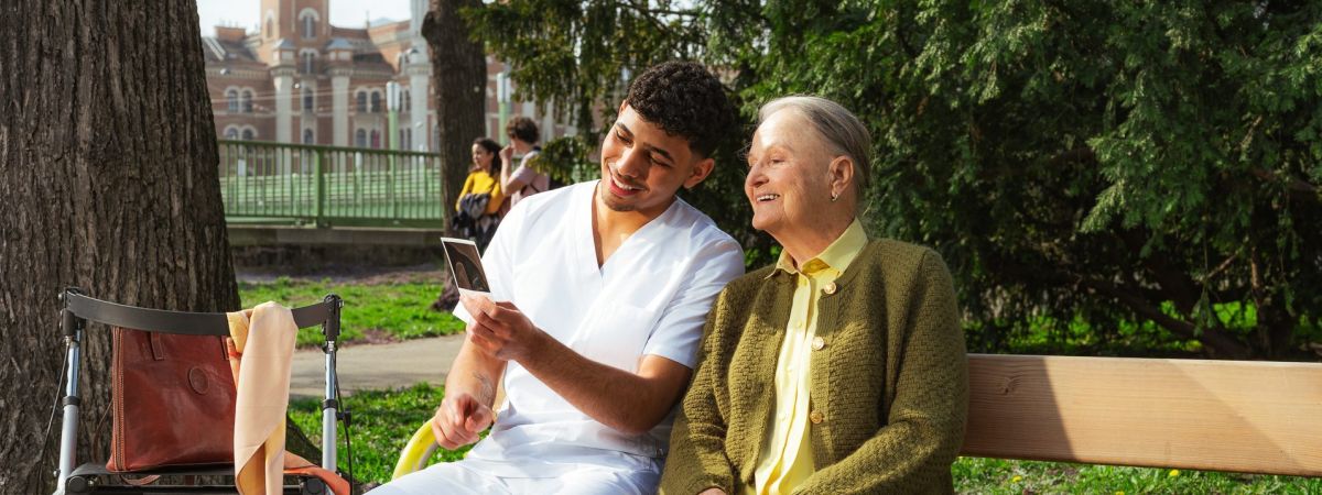 Eine ältere Dame und ein junger Pfleger sitzen im Park auf einer Bank und sehen sich gemeinsam ein Foto an.