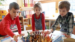 Drei Kinder stehen an einem Tisch mit Zeichnungen und Buntstiften.