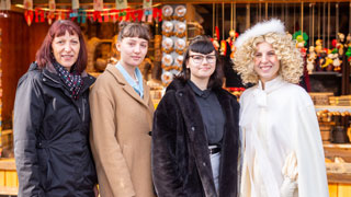 Direktorin, eine Schlerin als Christkind verkleidet sowie zwei Schlerinnen in Wintermnteln vor Adventmarkt-Stand
